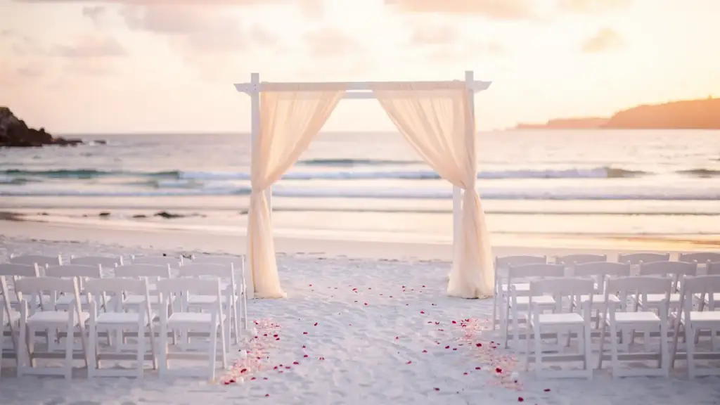 An empty wedding arch draped in sheer fabric on a beach at sunset, rows of chairs facing the sea